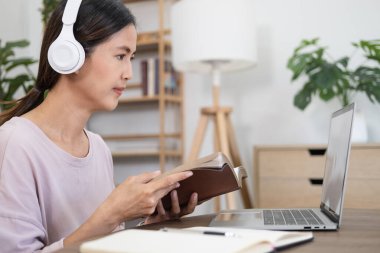 Sanal konferans. Kablosuz kulaklıklı genç bayanın portresi masada oturuyor, elinde kitap tutuyor ve okuyor, öğretmenle online ders alıyor ve dizüstü bilgisayara bakıyor. Evde çalışan bir kadın.