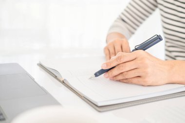 Günlük kıyafetlerle Asyalı iş kadını dizüstü bilgisayarla çalışırken not defterine notlar yazıyor, pencerenin önündeki temiz görüntüyü yumuşak ışıkla kapatıyor..