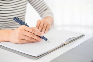 Günlük kıyafetlerle Asyalı iş kadını dizüstü bilgisayarla çalışırken not defterine notlar yazıyor, pencerenin önündeki temiz görüntüyü yumuşak ışıkla kapatıyor..
