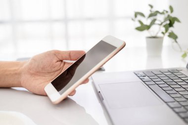 Asyalı iş kadını dizüstü bilgisayar kullanıyor ve akıllı telefonu kontrol ediyor, pencerenin önündeki temiz görüntüyü yumuşak ışıkta kapat.