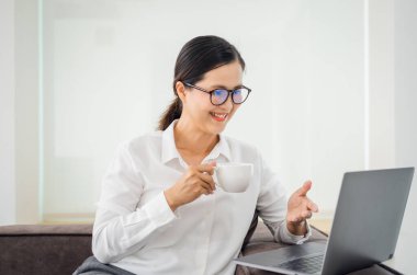 Ev ofisinin oturma odasında dizüstü bilgisayarda çalışan Asyalı olgun bir kadın. Uzaktan bilgisayar eğitimi, sanal eğitim, e-öğrenme kullanan Asyalı profesör evde online eğitim webinar izliyor.