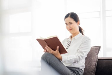 Evde oyalanalım. Milenyum Asyalı kadın rahat koltukta oturuyor, kapalı kapılar ardında. Güzel genç bayan heyecanlı hikayeler okuyor, tembelliğin tadını çıkarıyor, rahatlatıcı bir hafta sonu geçiriyor.
