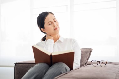 Evde oturma odasındaki kanepede kitap okuyarak fibromiyalji boynu ağrısından muzdarip yaşlı, stresli bir iş kadını. Yaşlı orta yaşlı kadın boyun masajı yapıyor. Okumaktan acı çekiyor..