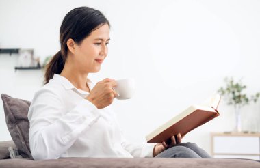 Evdeki oturma odasındaki kanepede kitap okuyan genç ve güzel bir kadın. Asyalı kadın tekstil kanepesinde oturmuş kahve içiyor. Beyaz duvar arkaplanı, kopyalama alanı, kapat.