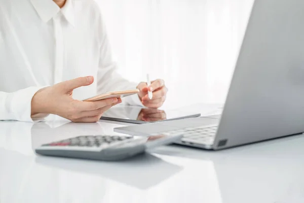 Beyaz elbiseli Asyalı iş kadını tablete not yazıyor ve akıllı telefonları kontrol ediyor. İş kadını, iş yerinde evraklar ve tabletlerle uğraşmakla meşgul. Temiz resmi kapat.