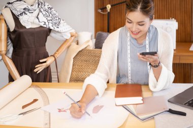 Genç, çekici Asyalı kadın moda tasarımcısı ofis masasına yaslanıyor, cep telefonuyla konuşuyor..