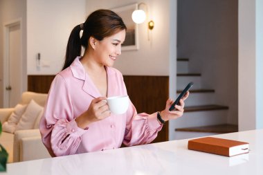 Genç Asyalı iş kadını ev telefonuyla kahve içiyor. Mutlu gülümseyen kadın profesyonel cep telefonuyla çalışıyor. Cep telefonundan mesaj atıyor..