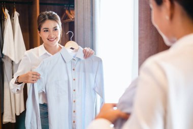 Elbise al, güzel Asyalı genç kadın, iş kadını tişört giyiyor, kadın giyiniyor işe gitmeden önce hazırlanıyor sabah evde aynada aynaya bakıyor..