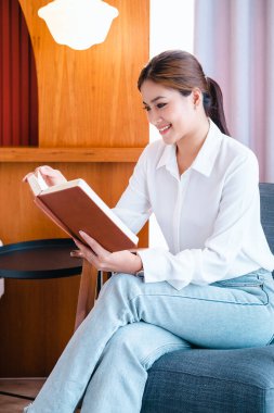 Güzel genç bayan heyecanlı hikayeler okuyor, tembelliğin tadını çıkarıyor, rahatlatıcı bir hafta sonu geçiriyor. Evde kalıp eğlenin. Milenyum Asyalı kadın rahat koltukta oturuyor, kapalı bir kitap gibi..