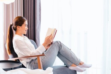 Asyalı kadın, sabah kalktıktan sonra pencerenin yanındaki koltukta kitap okuyor. Tembel Sabah yaşam tarzı konsepti. Heyecan verici hikayeler okuyan, rahatlatıcı bir hafta sonu geçiren güzel bir bayan.