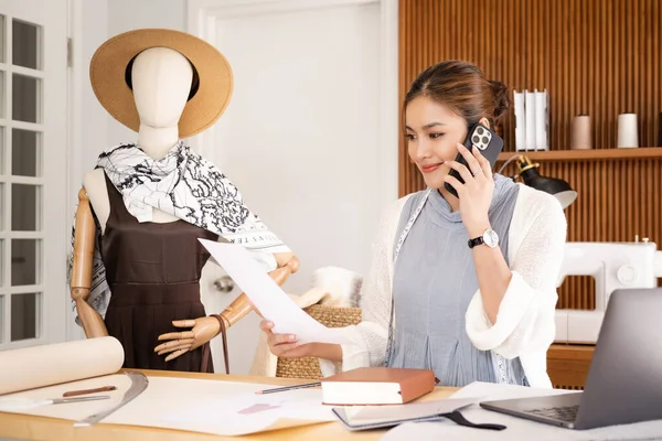 Mavi takım elbiseli, gülümseyen Asyalı genç faşist kadın moda tasarımcısının ofisinde oturuyor ve elinde telefon ve kağıt ile müşteriyle konuşuyor. .