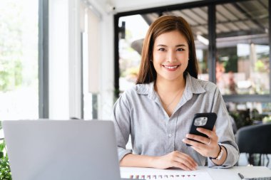 Modern ofisteki gülümseyen Asyalı kadın elinde cep telefonu, dizüstü bilgisayarının bebeğini tutuyor. 