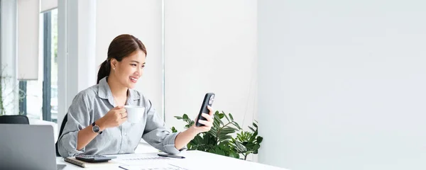 Beautiful cute asian young businesswoman in the co working space office cafe, using mobile phone videocall and drinking coffee smiling panorama copy space