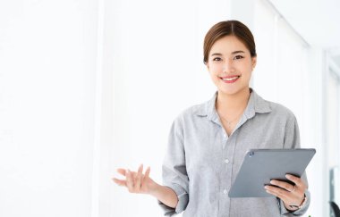 Genç Asyalı iş kadını tişört giyiyor, tablet kullanıyor ve modern çalışma alanında duruyor. Gülümseyen, kendine güvenen iş adamı kameraya bakıyor ve ofiste dikiliyor..