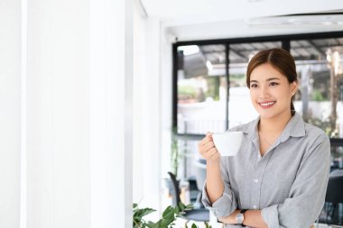 Genç iş kadını ayakta kahve içiyor ve modern ofisin penceresinden dışarı bakarken yeni bir başlangıç stratejisini düşünüyor. Boşluğu kopyala