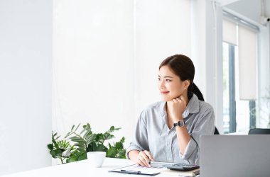 Çekici Asyalı iş kadını pencereden dışarı bakıp elinde kalemle ofiste dizüstü bilgisayarla çalışıyor. Boşluğu kopyala