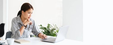 Gözlüklü Asyalı kadın dizüstü bilgisayara bakıyor, ödevini yapıyor, konsantre oluyor, çözüm arıyor ya da fikirler üretiyor. İş mücadelesi, görev konsepti üzerinde düşünme panorama afişi kopyalama alanı