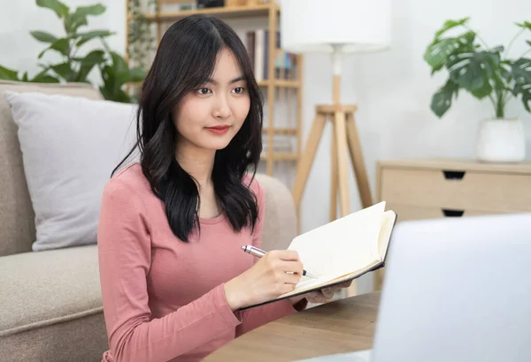 スタイリッシュなカジュアルな服を着た現代アジアの女性学生は リモートでラップトップを使用して勉強し オンラインレッスンを聞き リビングルームに座っている間 ノートブックでメモを取ります オンライン教育 — ストック写真