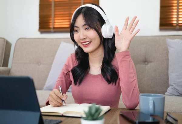 Asian woman  live online vlog say hi on virtual computer virtual home office. Young woman wearing head phone video call meeting on laptop alone at home. student taking notes while studying online.
