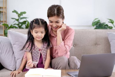 Kızı olan Asyalı bir aile anne yardımıyla tablet kullanarak ev ödevi yapıyor. Mutlu gülümse, Asyalı çocuk. Evde, oturma odasında otururken. Evde çevrimiçi öğrenim kavramı