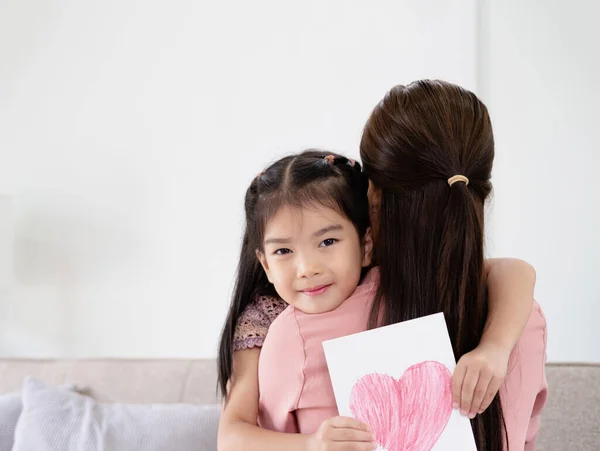 Smiling little girl holding greeting card for Happy Mother\'s Day with drawn red heart and hugging her mom. Loving asian family bonding together at home, closeup shot with free space