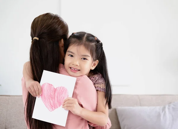 Congratulations. Loving asian daughter greeting surprised mom with Mother\'s Day, giving her handmade card and hugging, sitting on sofa at home interior, free space