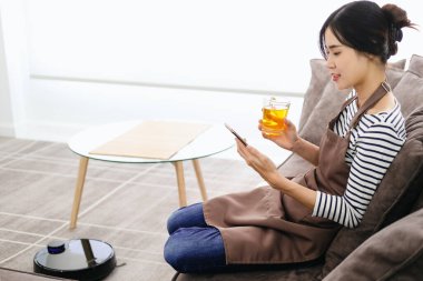 Kadın koltukta oturup dinlenirken siyah robotik süpürge yerleri temizliyor. Genç kadın yerleri temizlemek için otomatik süpürge kullanıyor, akıllı makine ev işi robotu kontrol ediyor..