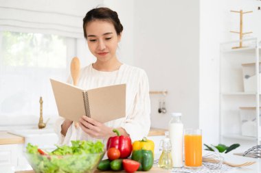 Gülümseyen ev hanımı yemek tarifi kitabına bakıyor evde hafif mutfakta sebze salatası pişiriyor. Sağlıklı yaşam tarzı konsepti.