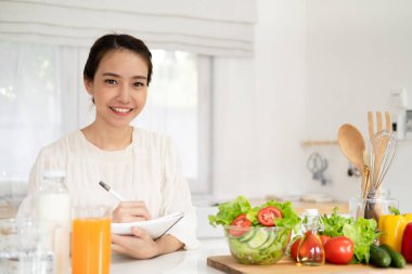 Mutlu gülümseyen Asyalı kadın masada salata suyu taze sebze ürünlerinin yanında deftere yazıyor. Keto diyeti