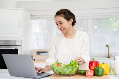 Mutfaktaki tablet dizüstü bilgisayarda yemek hazırlayan mutlu Asyalı kadın..