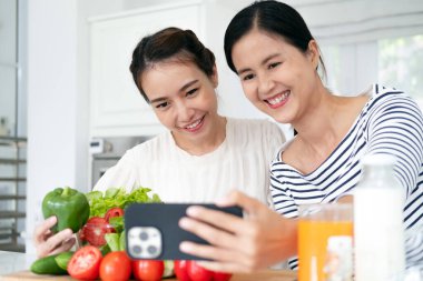 Önlük giymiş genç bir kadın mutfakta sağlıklı salata pişiriyor cep telefonuyla selfie çekiyor.