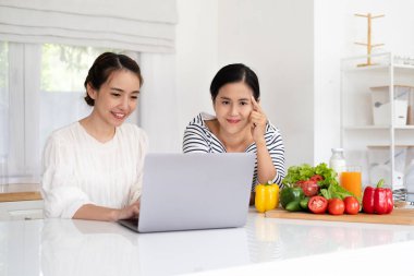 Neşeli çekici kız arkadaşlar mutfak masasının başında taze organik sebzelerle duruyorlar, modern bilgisayar kullanıyorlar, dizüstü bilgisayara bakıyorlar, internette güzel bir tarif arıyorlar,