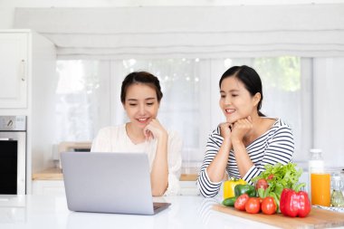 Neşeli çekici kız arkadaşlar mutfak masasının başında taze organik sebzelerle duruyorlar, modern bilgisayar kullanıyorlar, dizüstü bilgisayara bakıyorlar, internette güzel bir tarif arıyorlar,