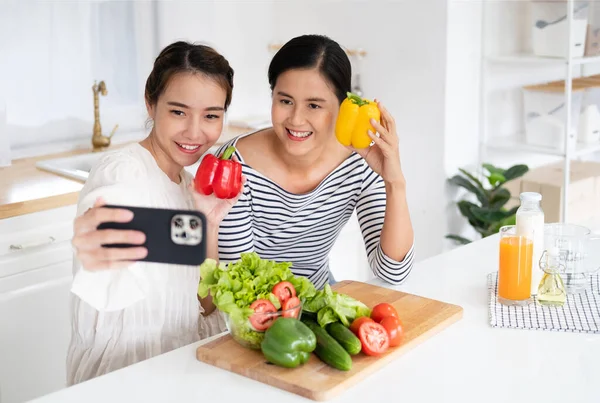 Önlük giymiş genç bir kadın mutfakta sağlıklı salata pişiriyor cep telefonuyla selfie çekiyor.