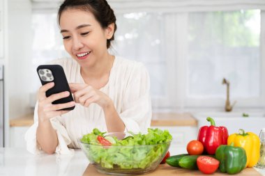 Asyalı bir kadın mutfak masasında içi taze sebzelerle dolu duruyor, ev hanımı elinde yemek uygulamaları, internet siteleri arama tarifleri, yemek hazırlama konusunda kafası karışmış arkadaşlarıyla sohbet ediyor.