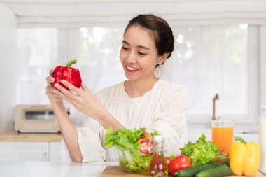 Sağlıklı yemek sağlıklı hayattır. Güzel genç Asyalı kadın elinde taze kırmızı dolma biber ve mutfakta sebze salatasıyla gülümsüyor.