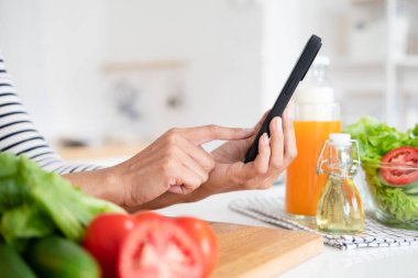 Mutfakta cep telefonuyla ellerinizi kapatın. İnternetten yemek yapmayı öğrenin. Genç bir kadın mutfakta yemek pişirme videosu seyrediyor. Yemek tarifini takip ederek salata yaparken yemek tarifi okuyor..