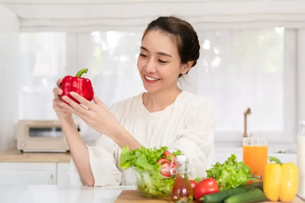 Healthy food is healthy life. beautiful young asian woman holding fresh red bell pepper and smiling with vegetable salad on table in the kitchen