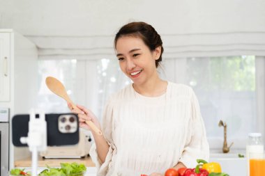 Profesyonel, güzel, mutlu Asyalı kadın mutfak kanalı için blogunda evinin mutfağında sağlıklı yaşamak ve tripodun üzerindeki kameraya bakmak hakkında yazıyor.