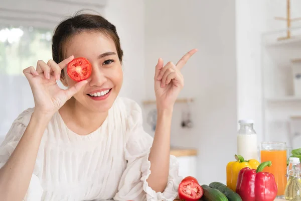 Young fun vegeterian housewife woman in casual clothes cover eye with tomato cooking food in light kitchen at home alone Healthy diet lifestyle concept.