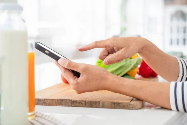 Close up of hands with mobile phone in kitchen.Learn cooking online. A young woman is watching cooking tutorial video in the kitchen.Reading recipe while making salad Following Recipe On phone.