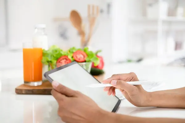 Close up hands holding pen pointing tablet in kitchen.Learn cooking online. woman is taking note in the kitchen.Reading recipe while making deit food Following Recipe On Digital Tablet.