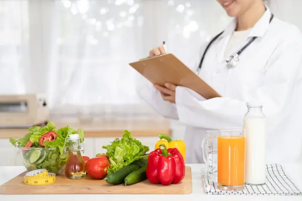 Woman dietitian in medical uniform with tape measure working on a diet plan sitting with different healthy food ingredients in  office on background. Weight loss and right nutrition concept