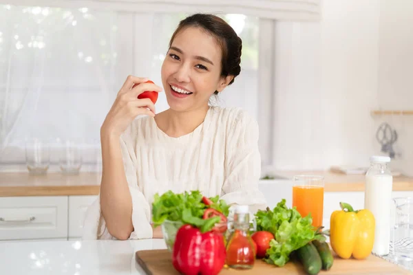 Healthy food is healthy life. beautiful young asian woman holding fresh red tomato and smiling with vegetable salad on table in the kitchen