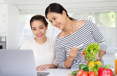 Neşeli çekici kız arkadaşlar mutfak masasında taze organik sebzelerle duruyorlar, modern bilgisayar kullanıyorlar, dizüstü bilgisayarı gösteriyorlar, internette güzel bir tarif arıyorlar,