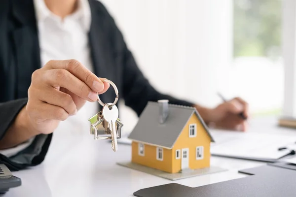 地产代理持有房屋钥匙的桌子上的房屋形状的钥匙圈与房屋设计文件 计算器 模型房子 投资财产的概念 免版税图库图片