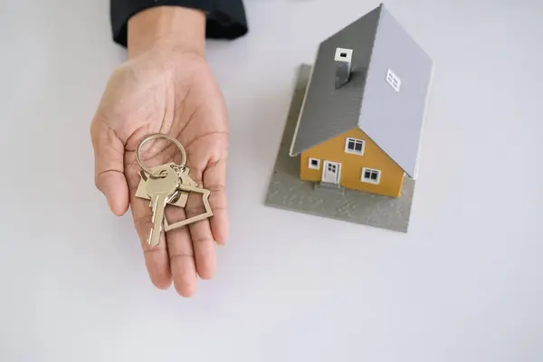 地产代理持有房屋钥匙的桌子上的房屋形状的钥匙圈与房屋设计文件 计算器 模型房子 投资财产的概念 图库图片