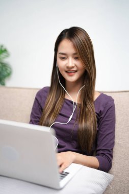 Kulaklıklı genç Asyalı kadın. Yazıyor ve not alıyor. İnternetteki laptop ekran çalışmasına bakın. Kulaklıklı mutlu kadın bilgisayarla uzaktan çalışıyor. Eğitim kavramı.