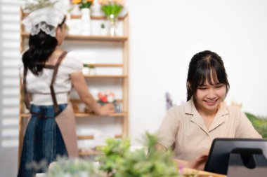 Küçük işletme, ev dekorasyonu konsepti. Etrafı çiçeklerle çevrili Asyalı kadın çiçekçi, küçük bir çiçekçi dükkanı sahibi, tablet ya da cep telefonu kullanıyor. .