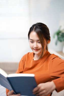 Kız, oturma odasında ilginç bir roman okumaktan zevk alıyor. Hobiler ve aktiviteler. Mutlu Asyalı kadın kitap okuyor, kanepede oturuyor ilginç bir hikayenin tadını çıkarıyor, sınava çalışıyor.,
